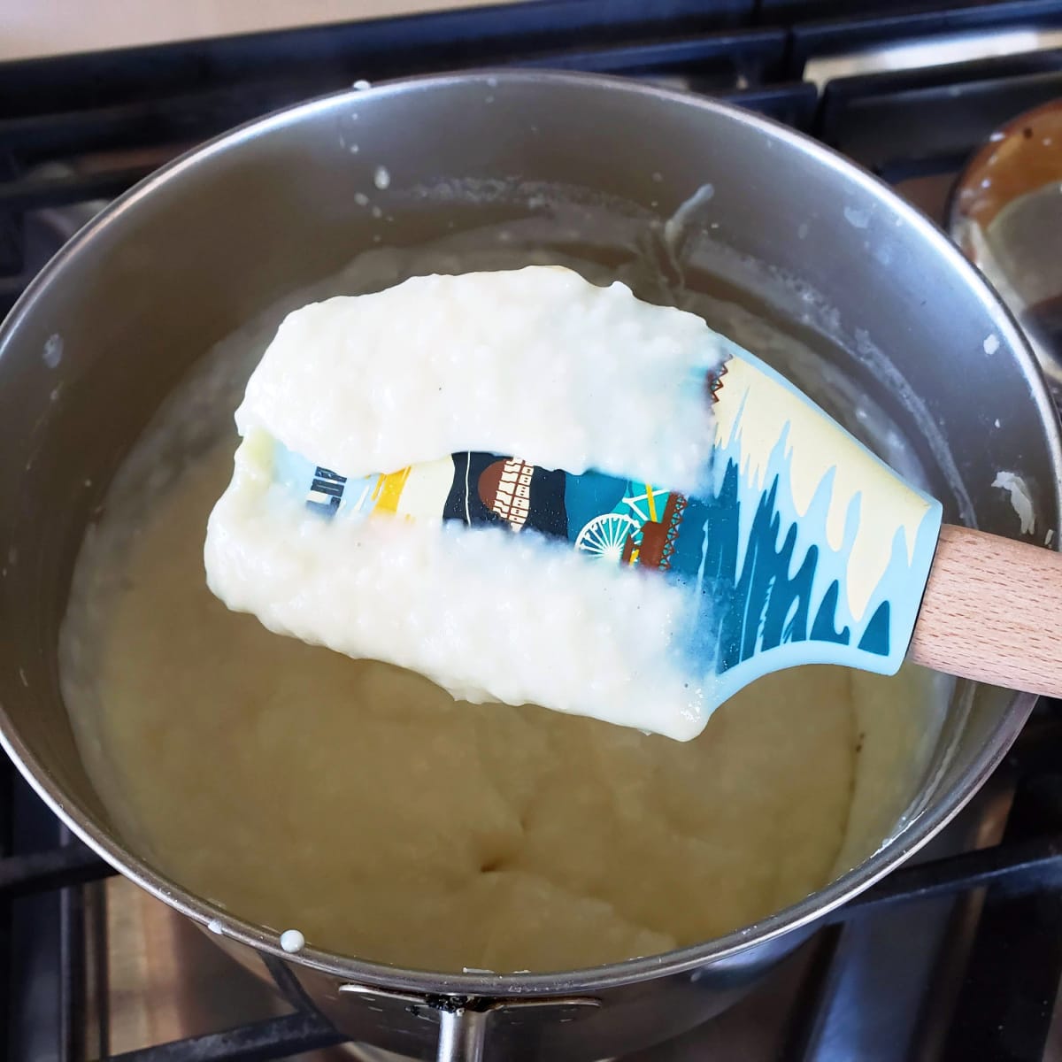 A colorful spatula has white coconut pudding on it held above a pot of the pudding on ShockinglyDelicious.com
