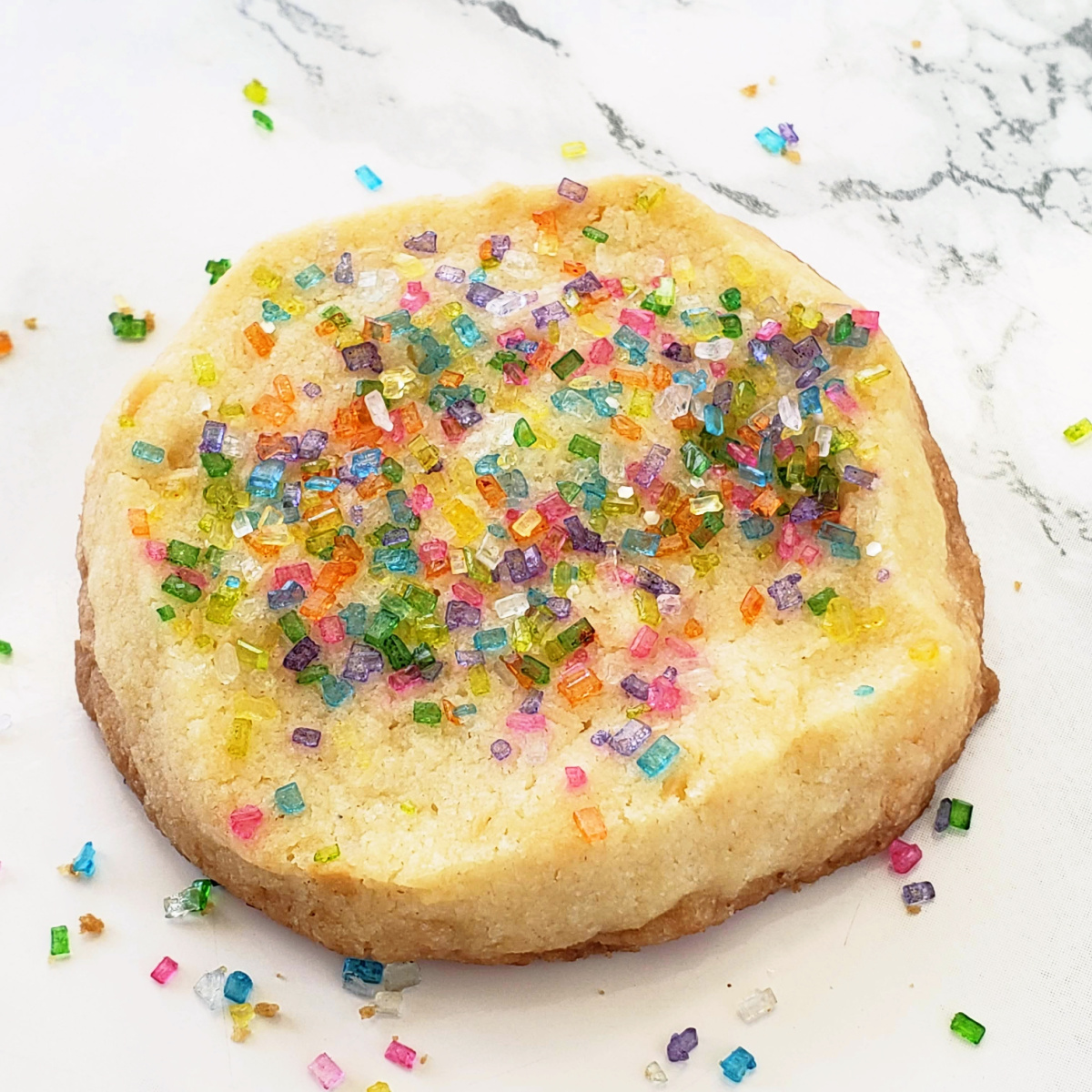 Yellowish cookie with colored sugar crystals on top sits on a white marble counter on ShockinglyDelicious.com