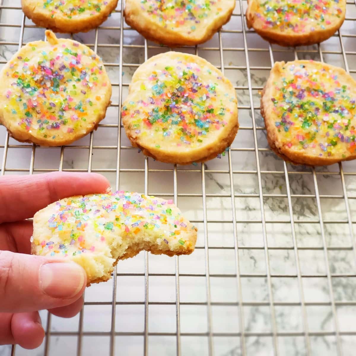 A hand holds a bitten cookie with other cookies in the background on a cooling rack
