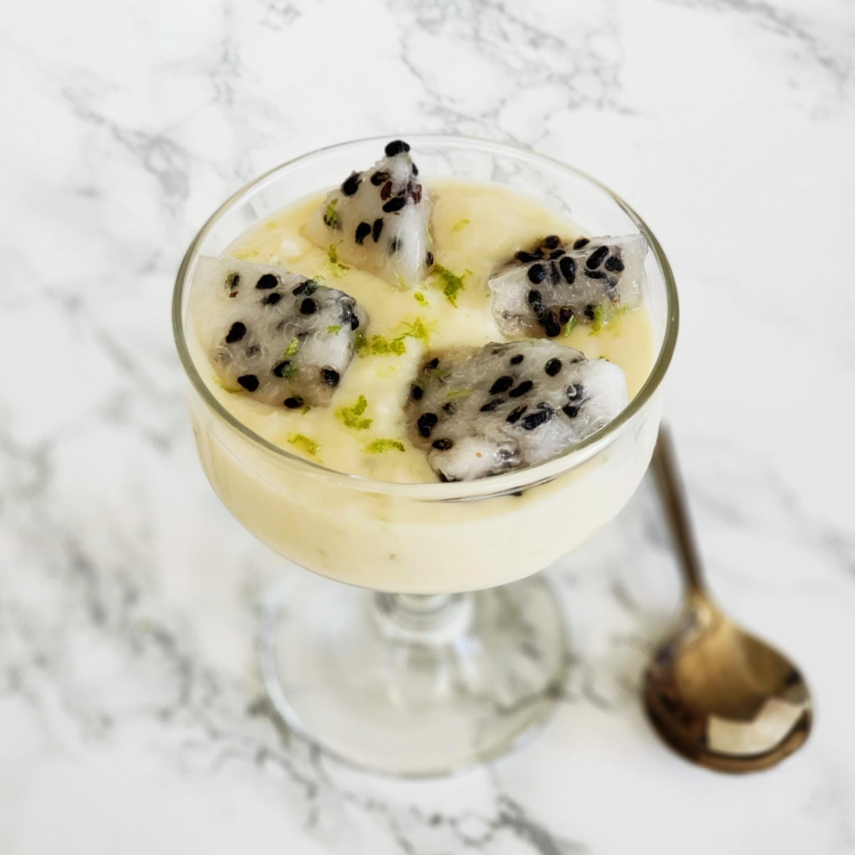 Coconut Pudding in a glass goblet with 4 pieces of white dragon fruit on top, a spoon alongside, on a white marble countertop