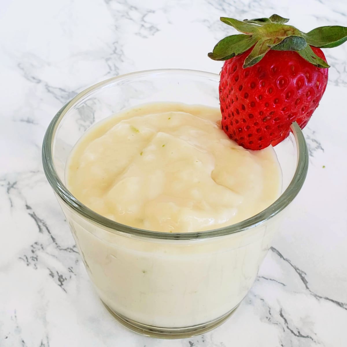 Coconut Pudding in a glass cup with a strawberry perched on the edge, on a white marble counter
