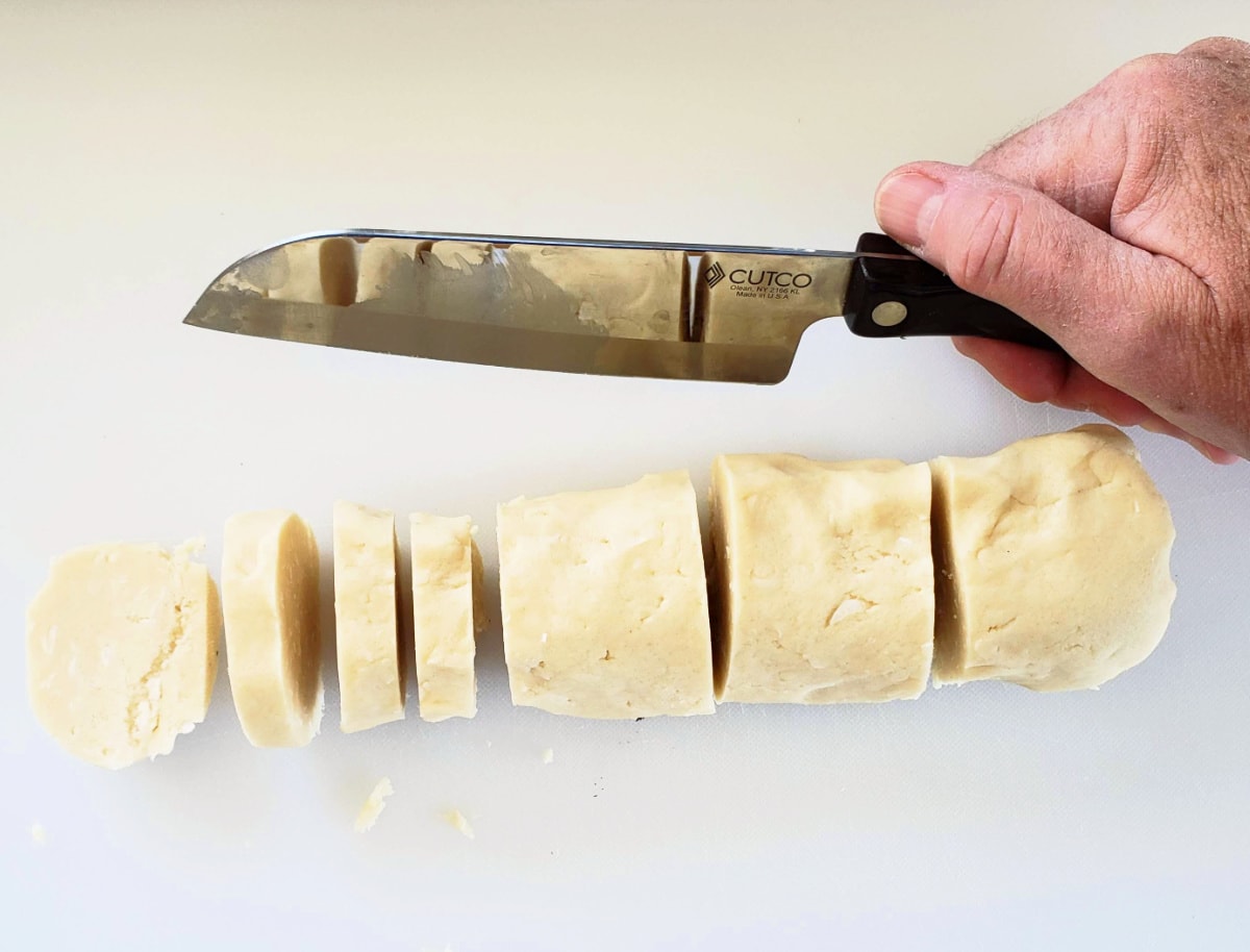Hand holding knife next to sliced log of cookie dough on a white cutting board