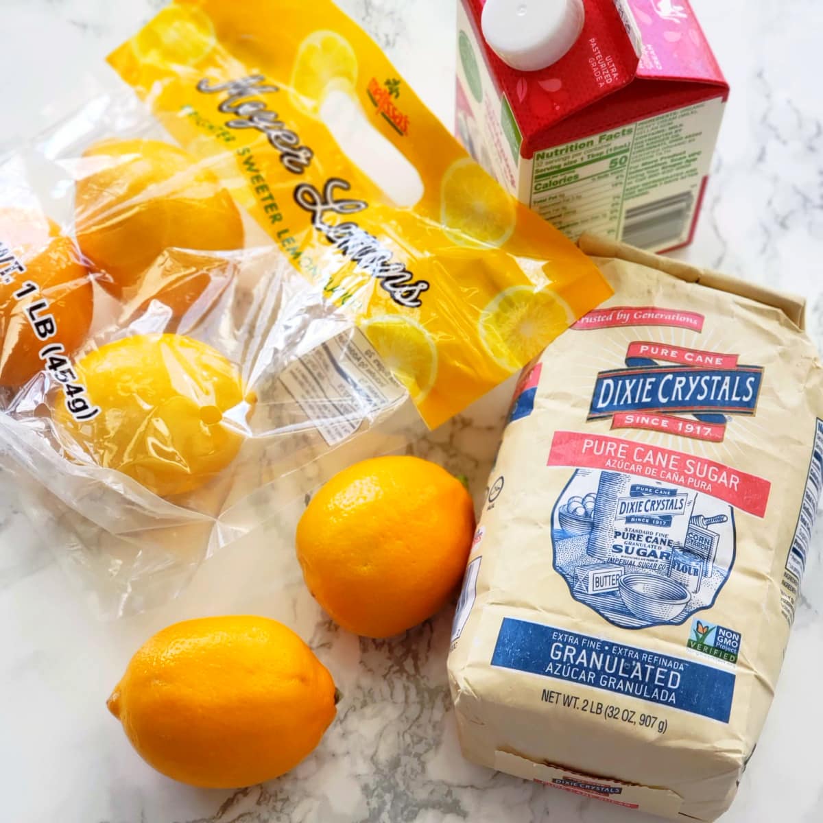 A bag of sugar, lemons and a carton of cream are laid on a white marble counter to make Meyer Lemon Posset on ShockinglyDelicious.com