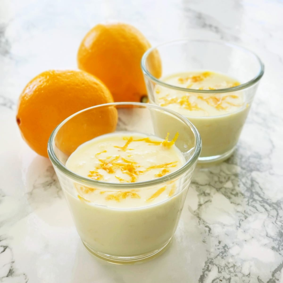 2 glass cups of yellow dessert with lemon zest on top with 2 lemons behind them on a white marble counter