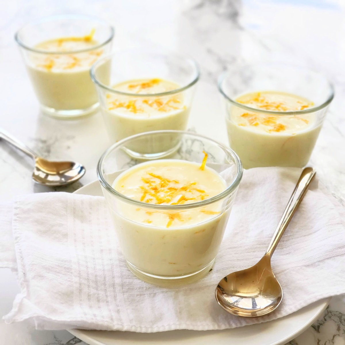 4 glass cups of Meyer Lemon Posset with 2 spoons alongside, on a white marble counter with a white napkin