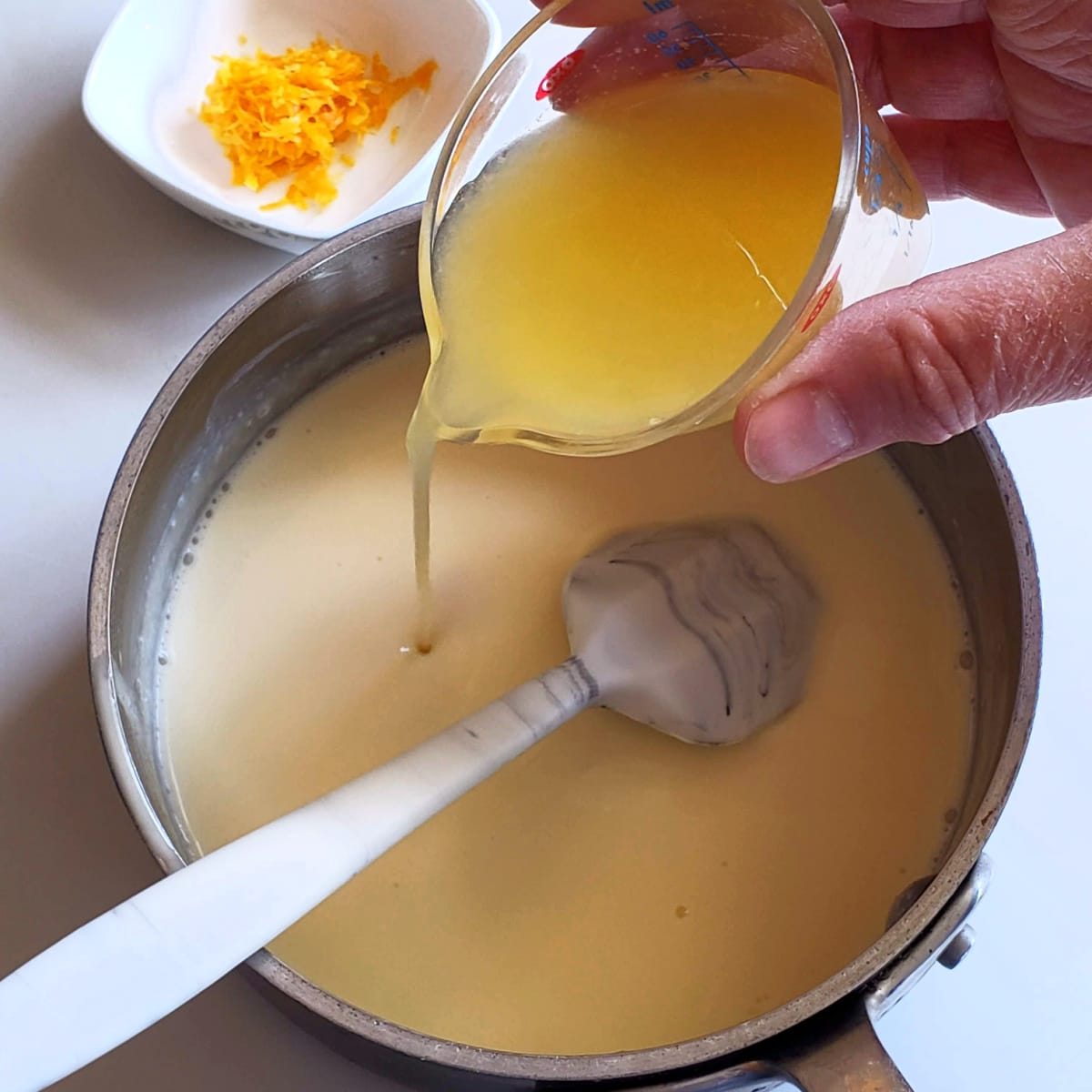 A hand pours lemon juice from a small measuring cup into liquid in a silver pan, with a white bowl of lemon zest in the background