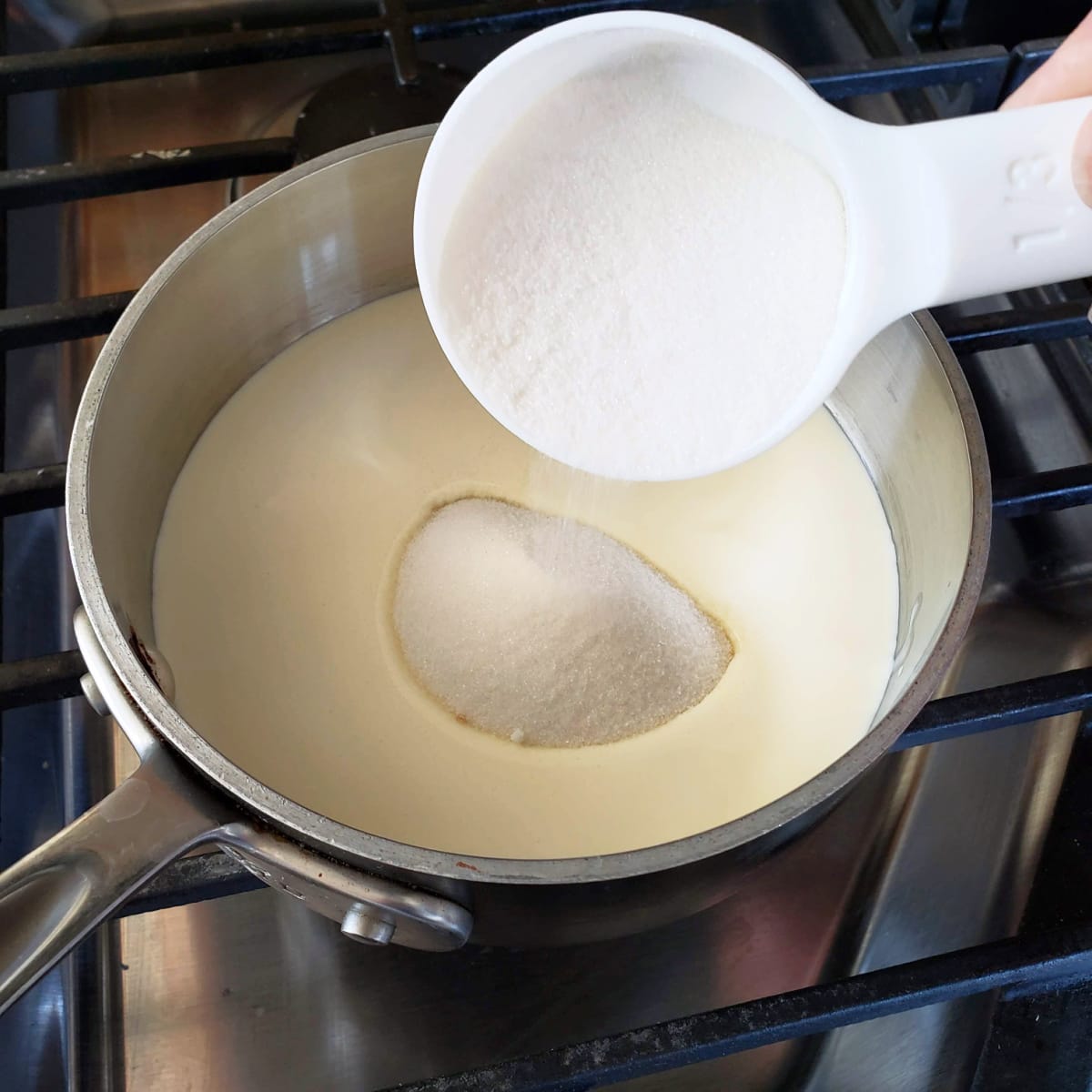 White sugar being poured into a small silver saucepan with cream, sitting on a stovetop