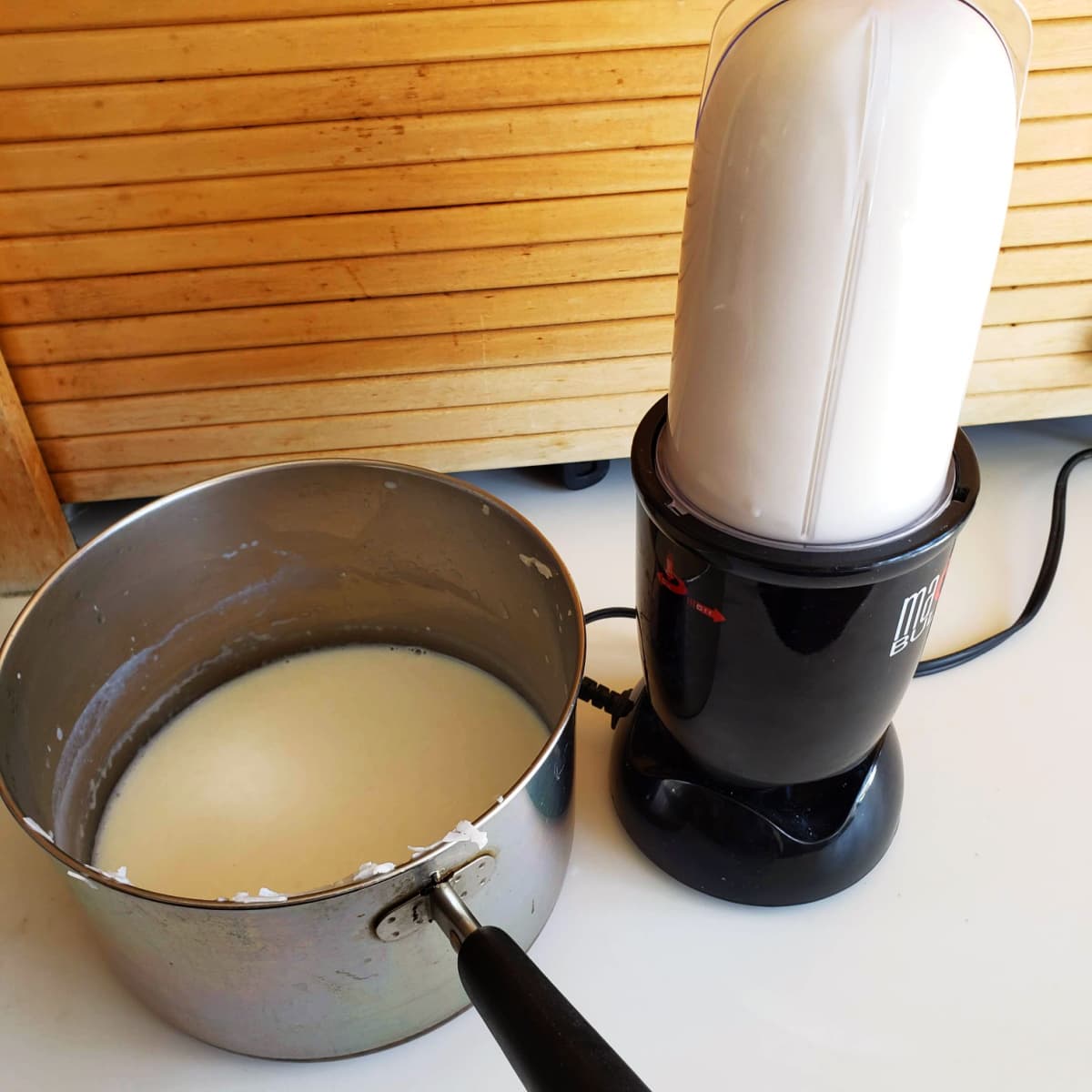 A pot with milk in it sits next to a small blender with a white mixture in it, in front of a wooden background