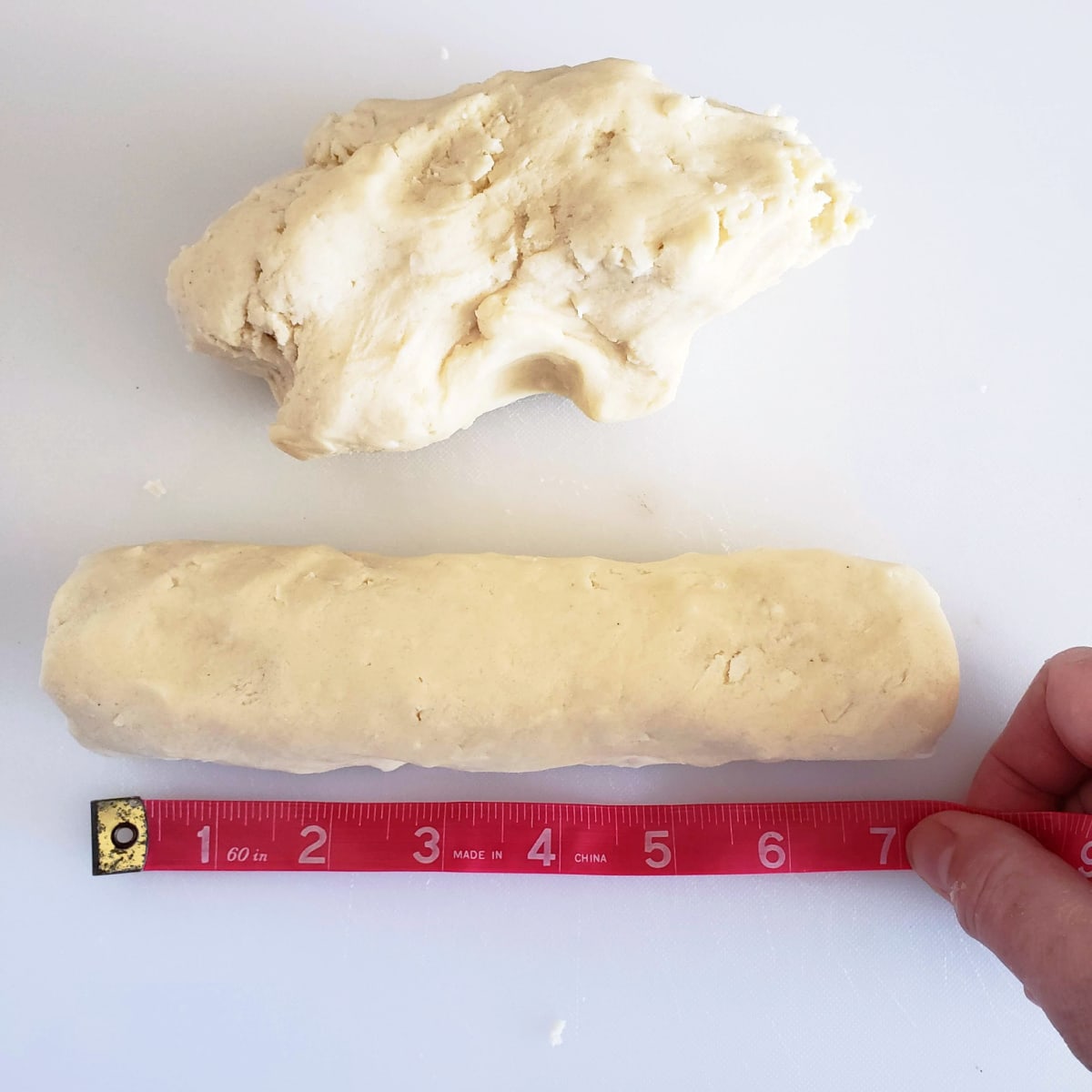 1 roll of Coconut Lime Shortbread dough is being measured by a pink tape measure, while a pile of unshaped dough rests behind it on a white cutting board