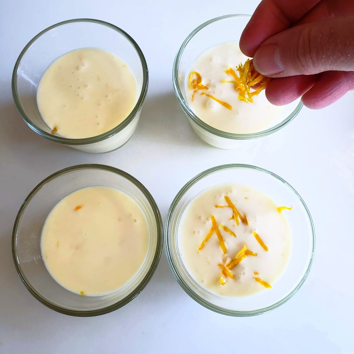 A hand sprinkles lemon zest on top of a glass with Meyer Lemon Posset in it next to 3 other glasses on a white background