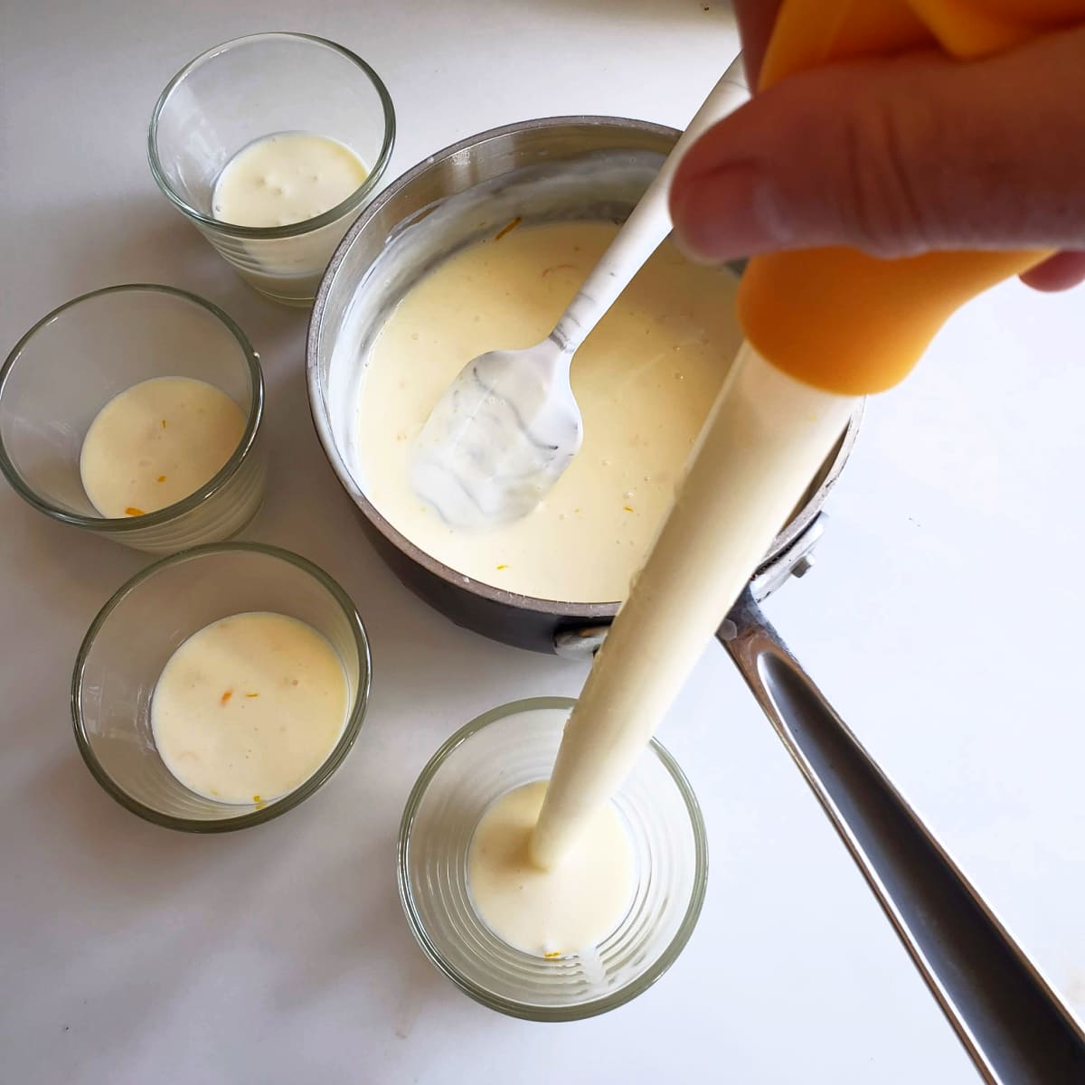 A hand holds a baster filled with liquid being transferred into 4 small glasses alongside a pot containing the rest of the liquid