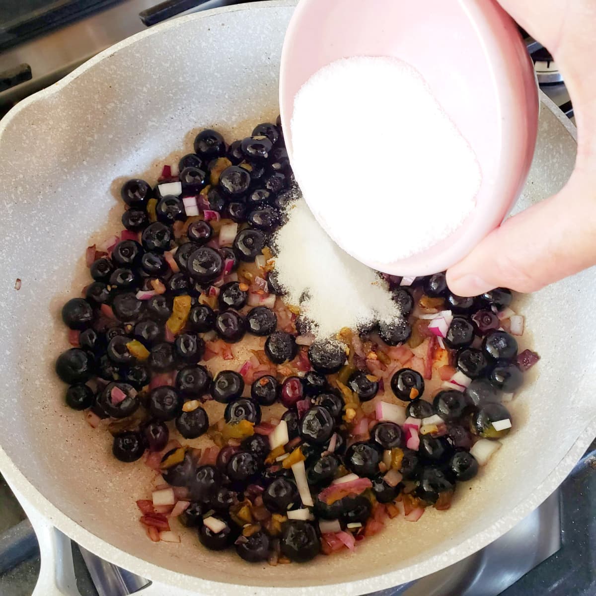 Hand holds small pink bowl pouring sugar into white skillet with blueberries