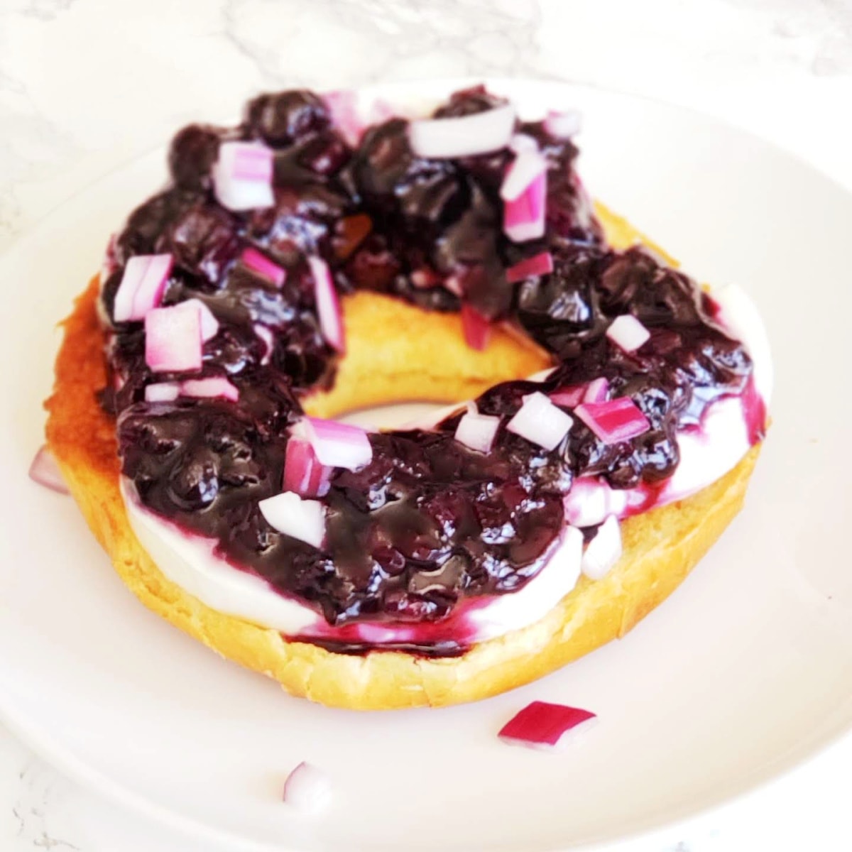 Blueberry Hatch Chile Chutney with chopped purple onions garnishing, on top of a toasted bagel half on a white plate with white marble countertop