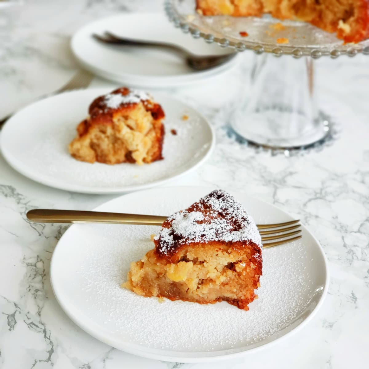 2 wedges of Apple Almond Olive Oil Cake on white plates with forks, with remaining cake on a glass cakestand behind