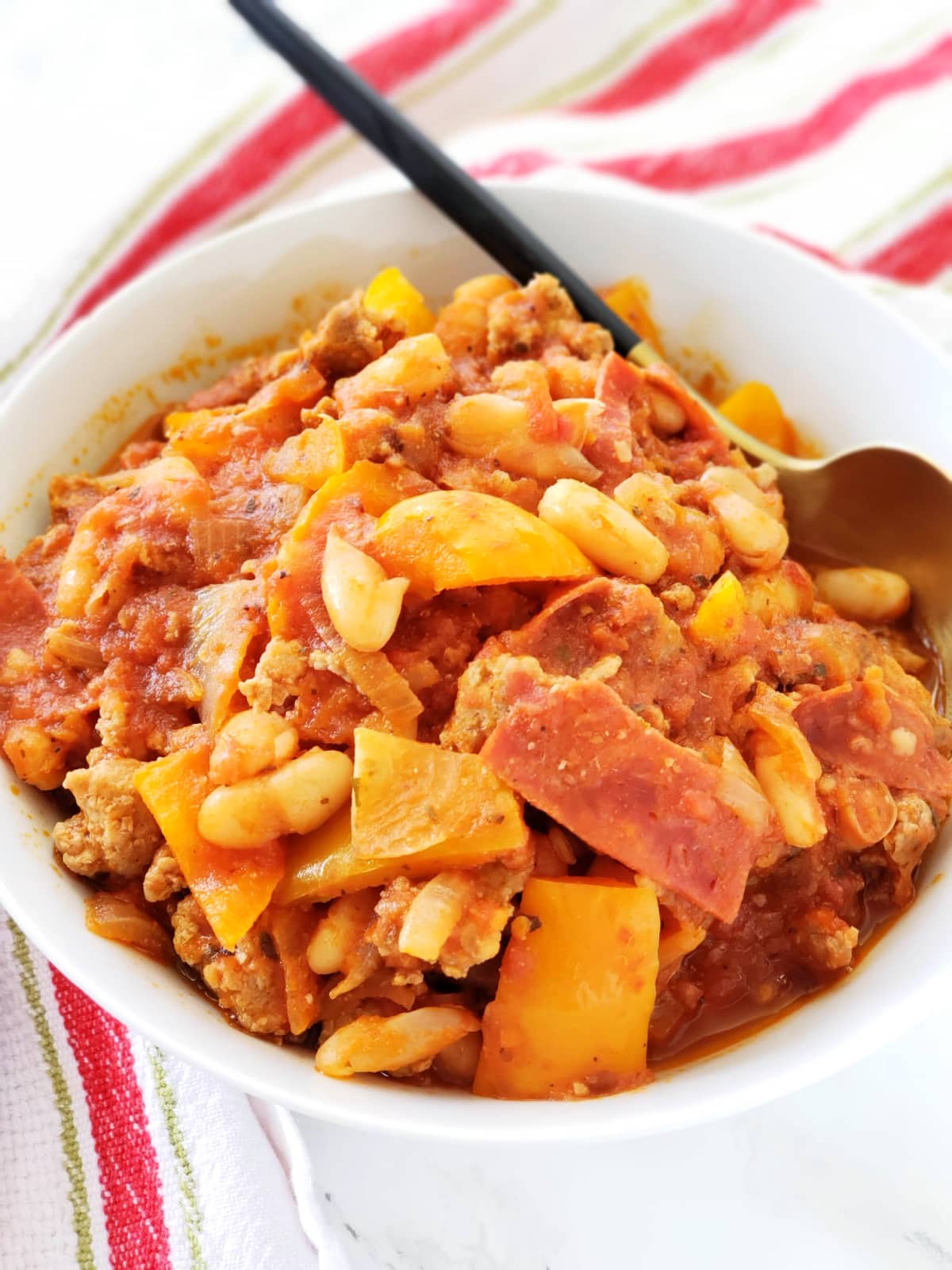 Pizza Chili in a white bowl with a spoon nestled in the bowl, atop a red striped towel on ShockinglyDelicious.com