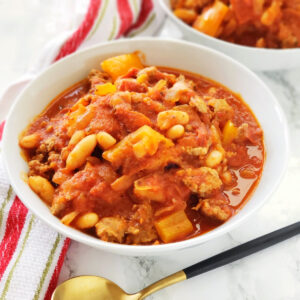 White bowl of Pizza Chili on a red and white striped dish towel on a white counter with a spoon in front