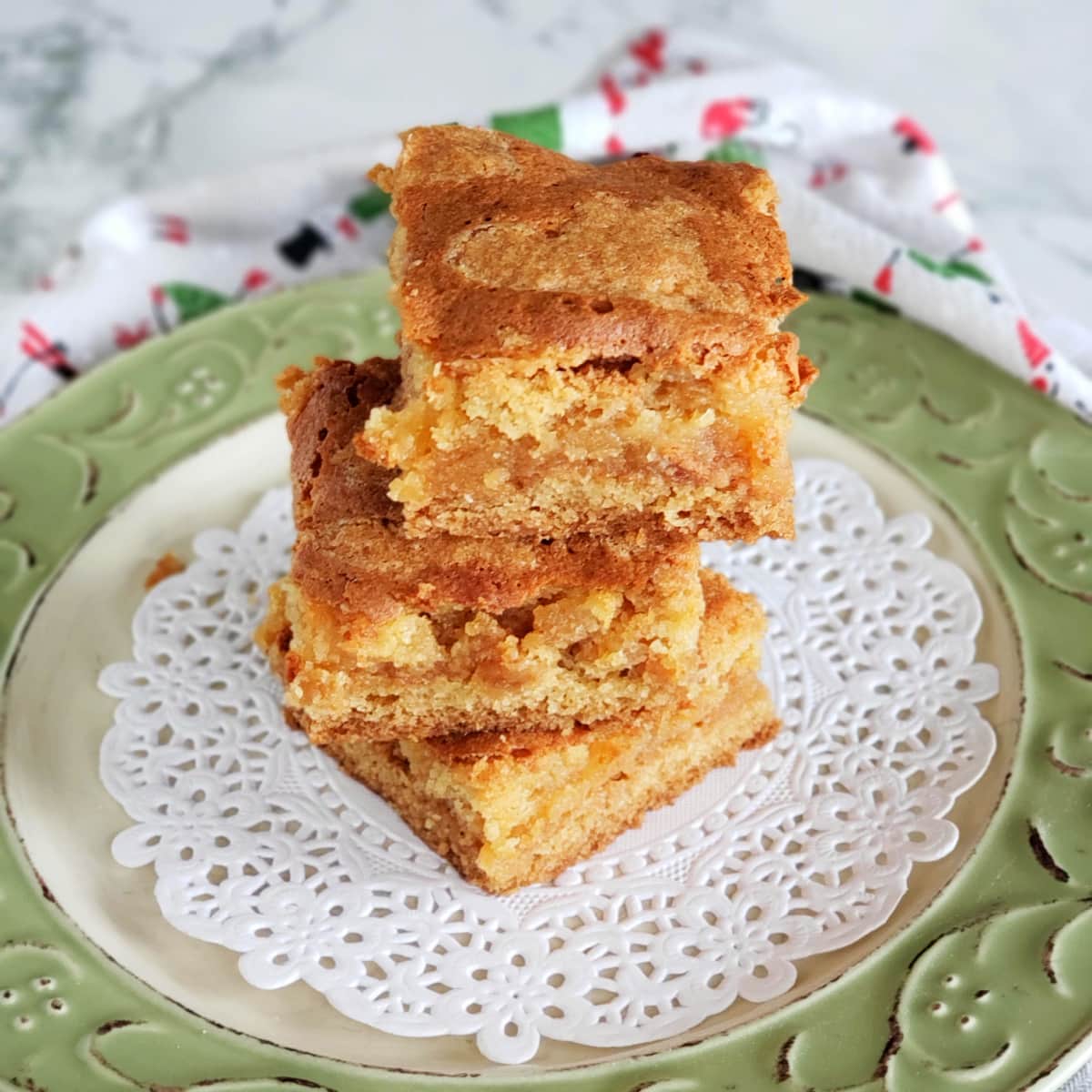 3 brown bar cookies stacked on top of each other on a green-rimmed plate with a doily in the center, on top of a Christmas towel