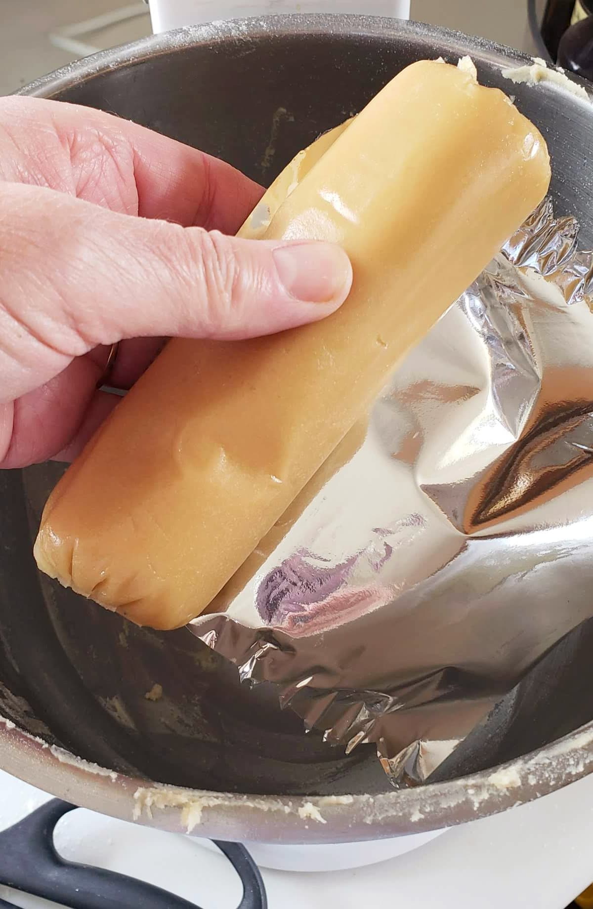 A hand holds a thick cylinder of almond paste removed from its shiny silver wrapper