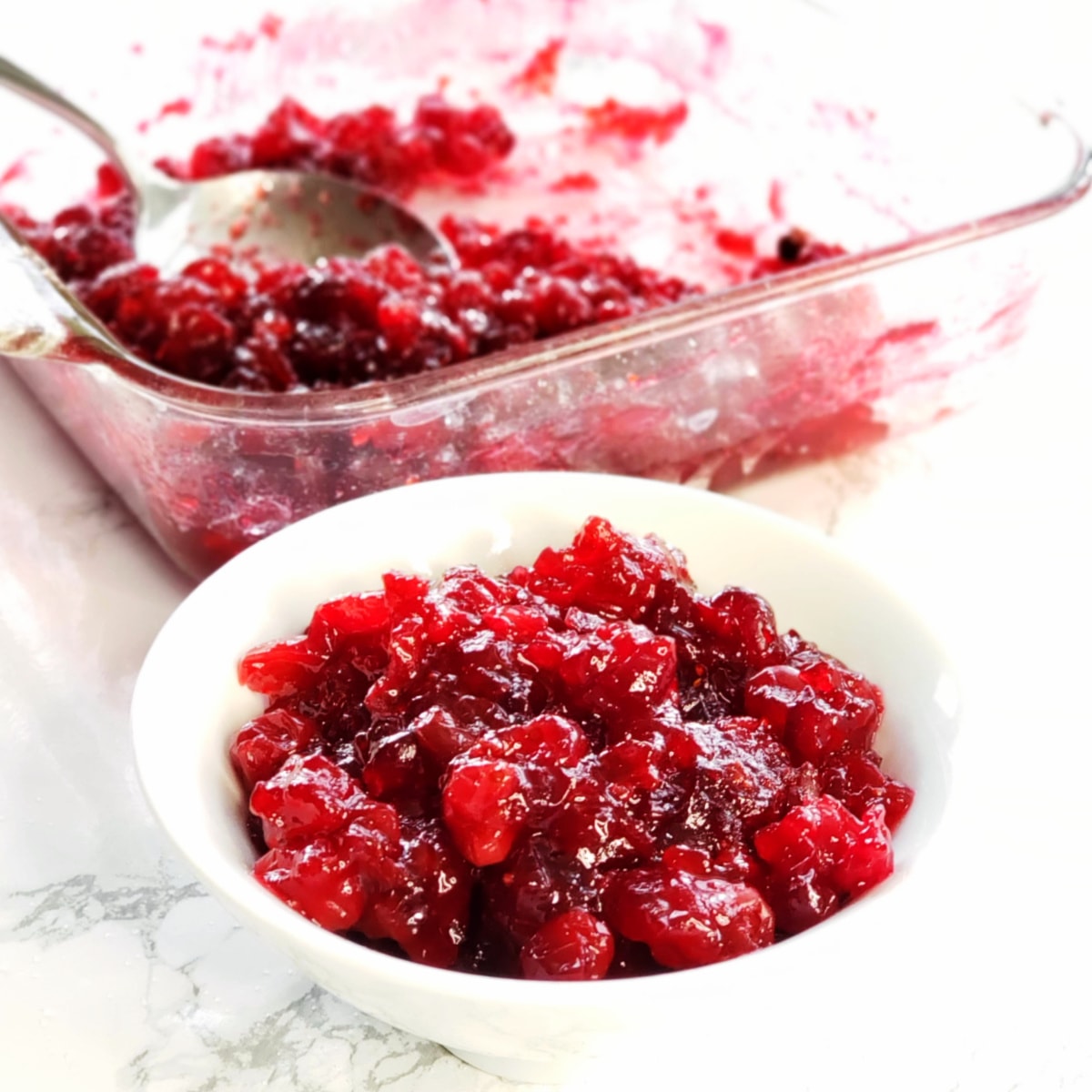 Baked Cranberry Sauce with Orange Liqueur in a white bowl with a clear glass baking dish of cranberries in the background