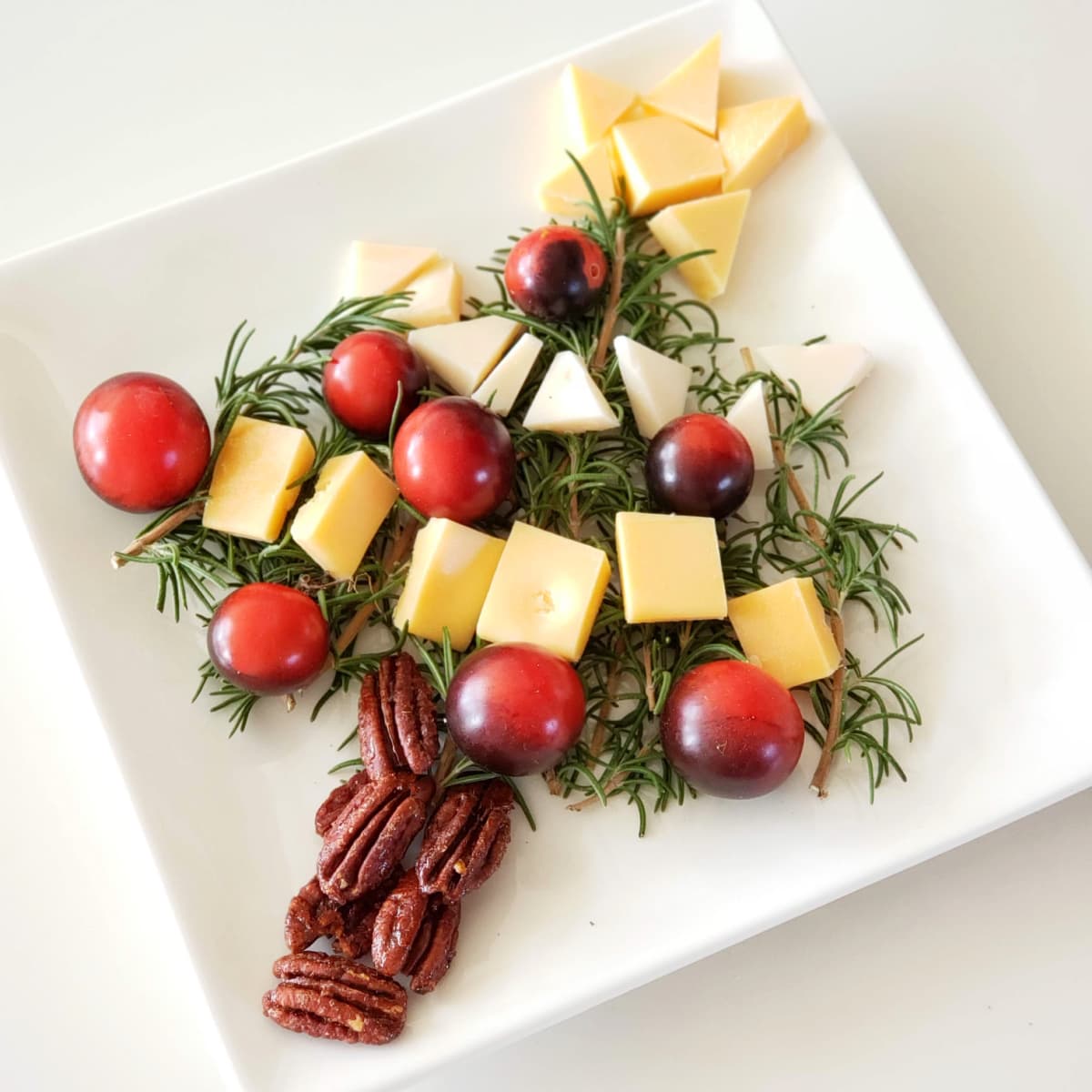 Christmas tree charcuterie plate with rosemary for the leaves and branches and cheese and cherry tomatoes for the ornaments, with Maple Glazed Pecans as the tree trunk