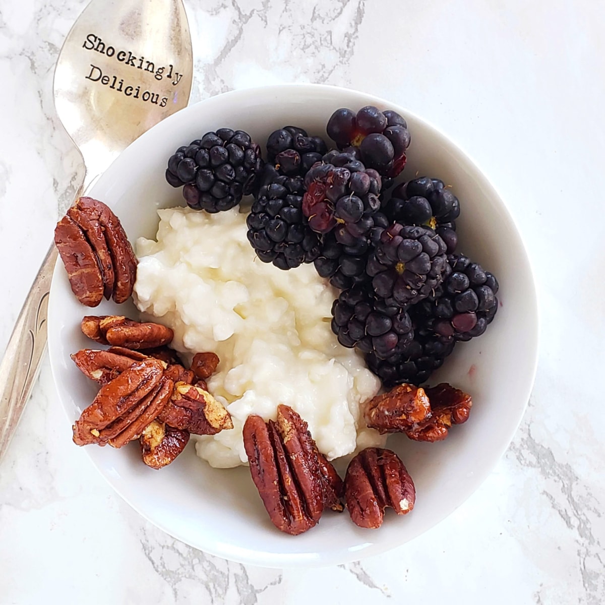 White bowl with cottage cheese, blackberries and glazed pecans on top, with a silver spoon alongside, sitting on a white marble counter