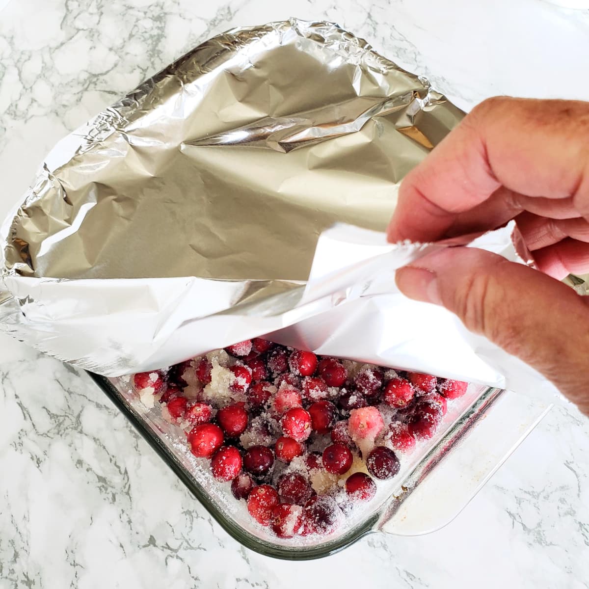 A hand covers baking dish with cranberries in it with foil