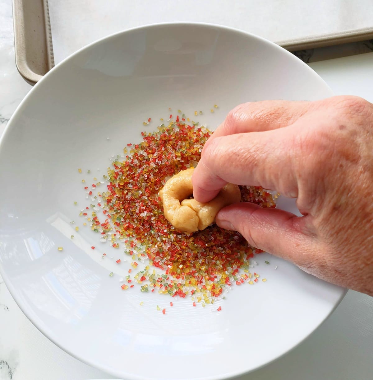 Hand holds a donut-shaped cookie that is dipped into colored sugar crystals in a white bowl