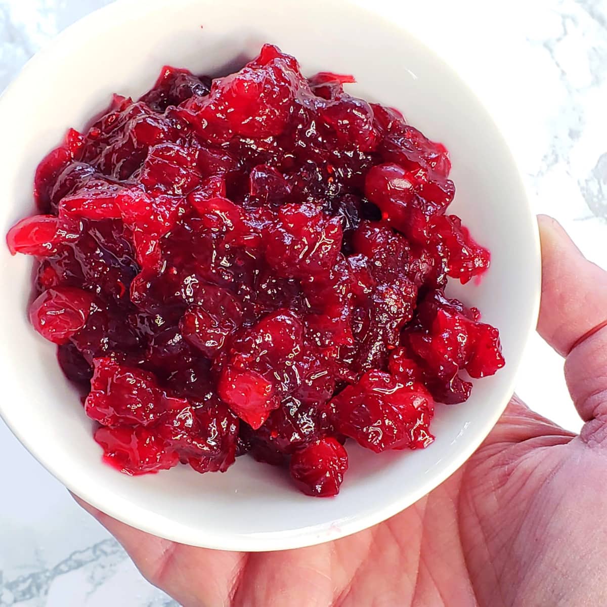 Easy Baked Cranberry Sauce with Orange Liqueur in a white bowl held by a hand against a white marble countertop