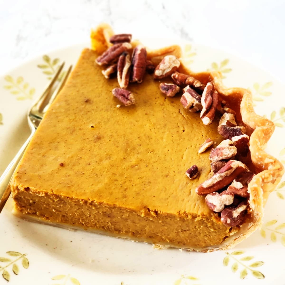 Wedge of Eggnog Pumpkin Pie with an outer rim of pecans on a beige plate with tree-decorated outer rim on ShockinglyDelicious.com