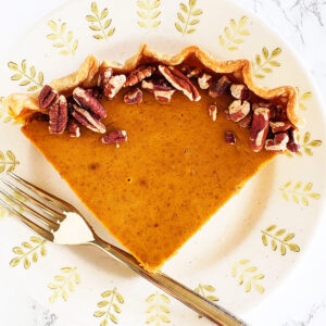 A wedge of Eggnog Pumpkin Pie on a beige plate with a rim decorated with green tree shapes, and a fork on the left side of the plate