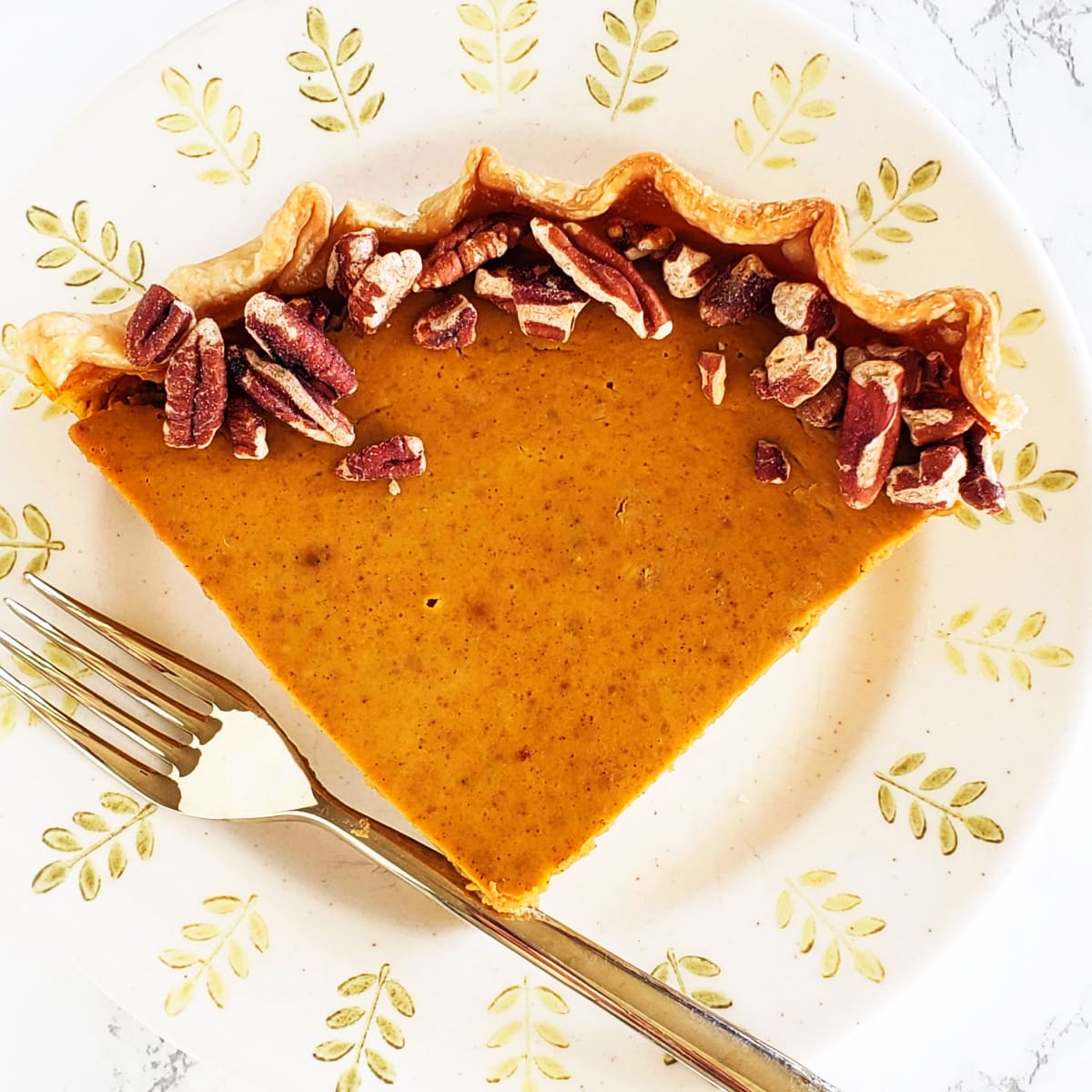 A wedge of pumpkin eggnog pie on a beige plate with a rim decorated by green trees, and a fork on the left side of the plate