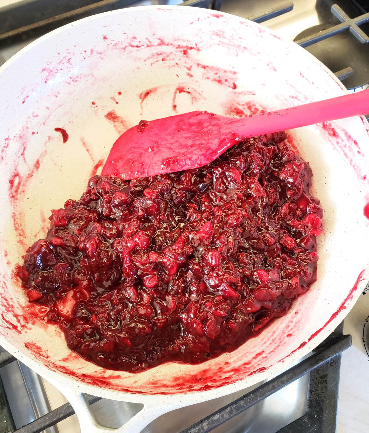 A red spatula in a white skillet shows how thick and jam-like the cranberry sauce isd jam-like