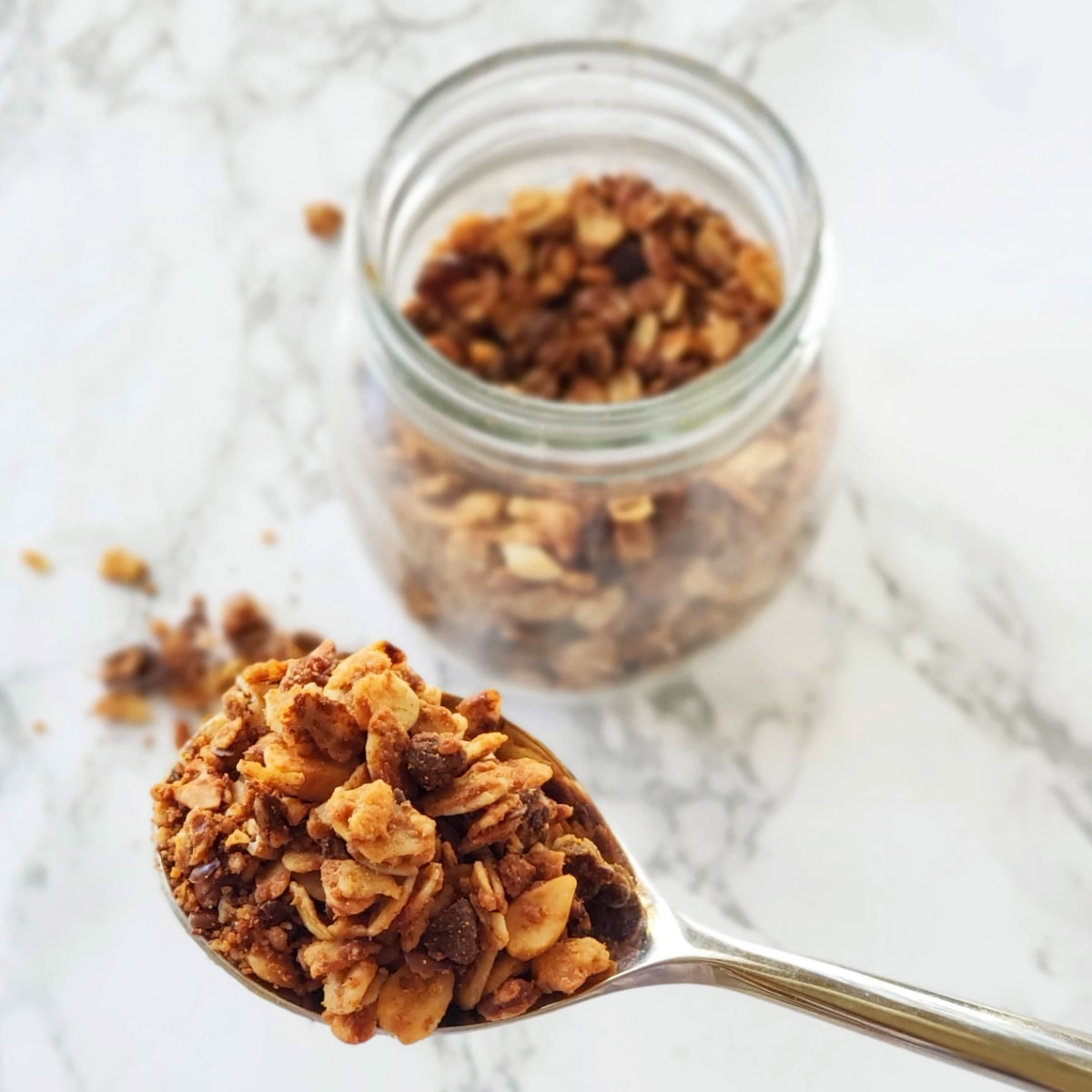 A spoon with granola in it in foreground and a clear glass jar of granola in background on a white marble counter