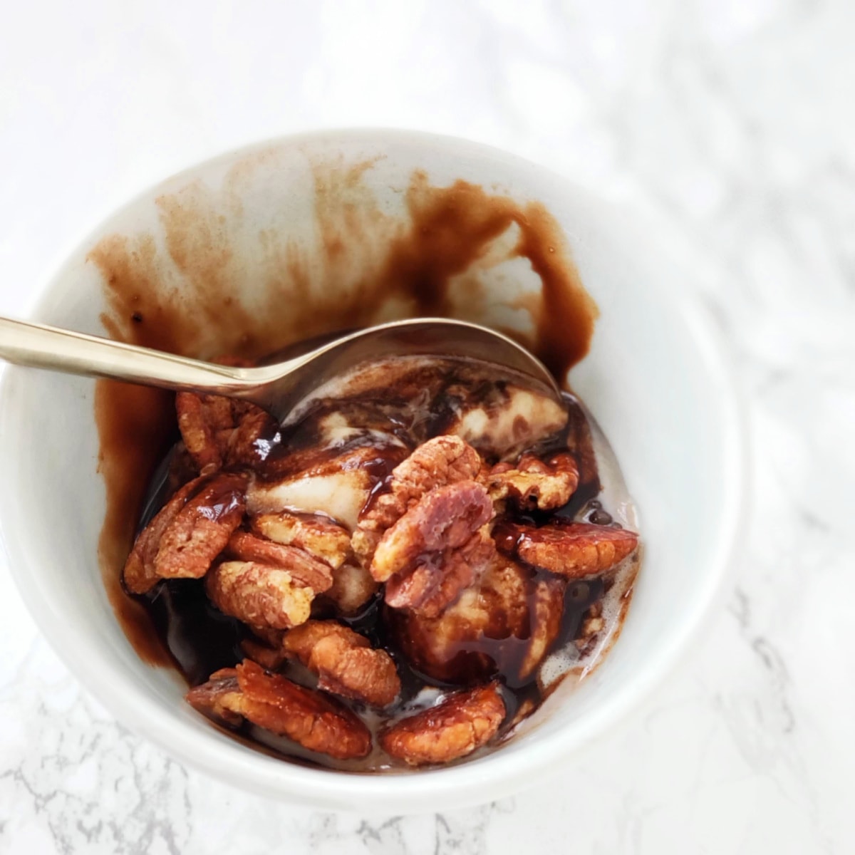 Vanilla ice cream topped with chocolate sauce and pecans in a white bowl with a spoon in the bowl