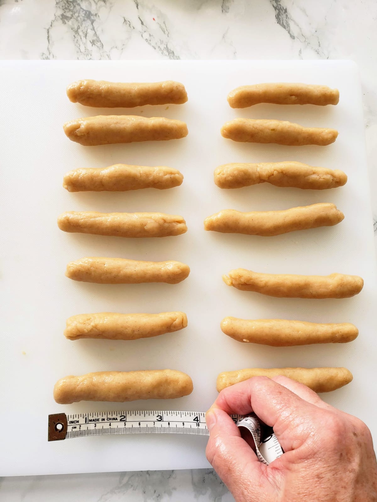 2 columns of 7 cylinders of rolled cookie dough on a white cutting board, with a hand holding a measuring tape to measure their length