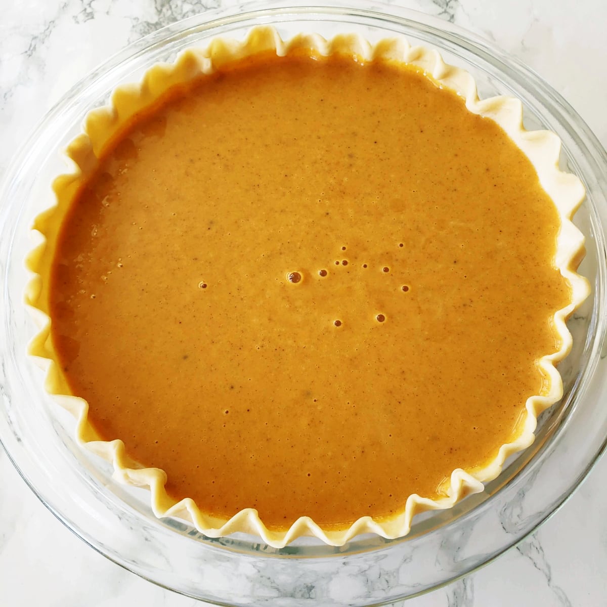 Brown colored pie filling in pie shell in a clear glass pie dish ready to bake for Eggnog Pumpkin Pie