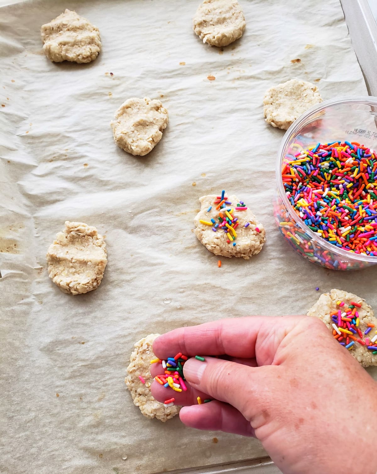 Cookies on parchment paper with a handle adding sprinkles and a container of sprinkles in the backgroundrs to flatten cookies and add sprinkles on top