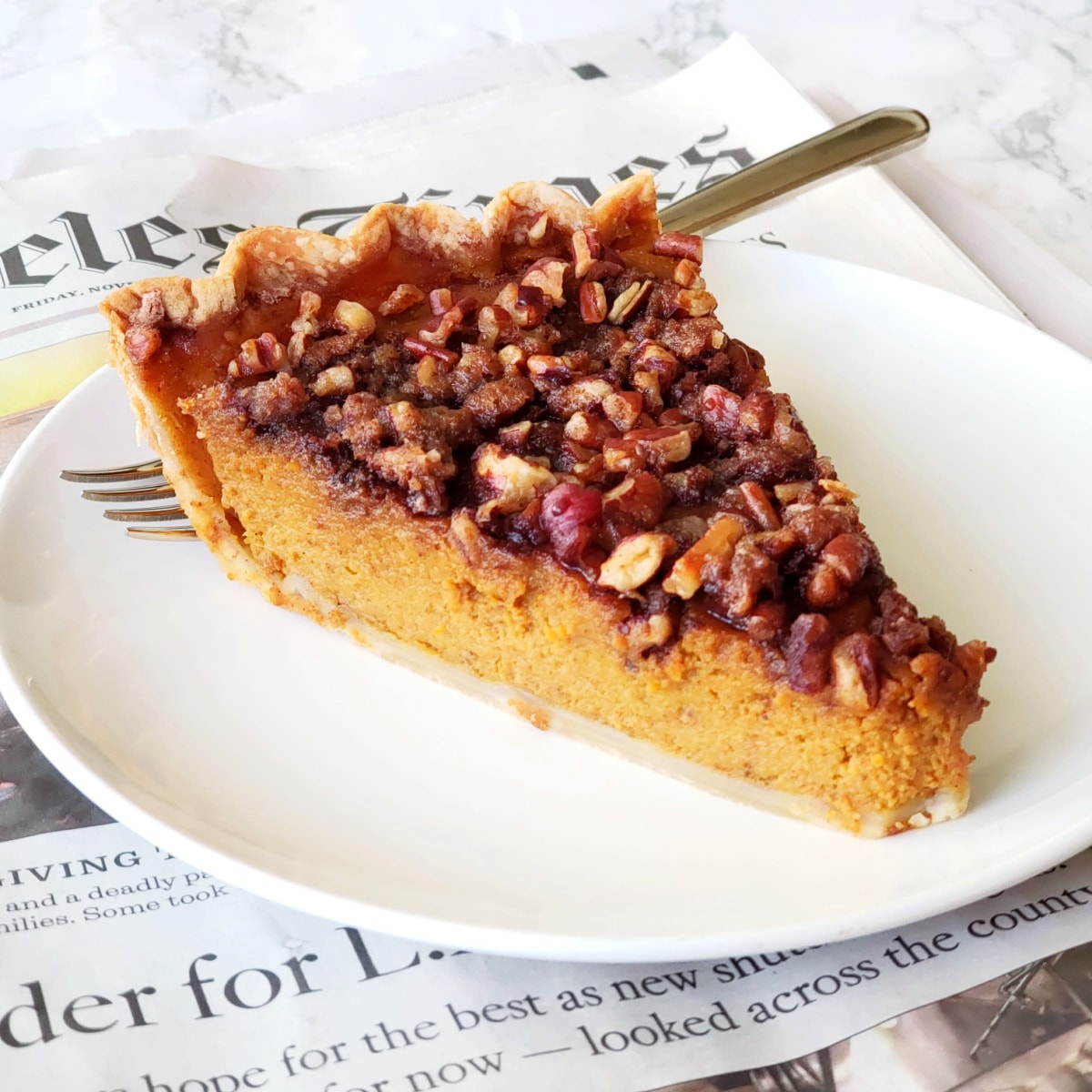 Slice of pumpkin Pie with nuts on top on a white plate with fork behind, sitting on a copy of the newspaper