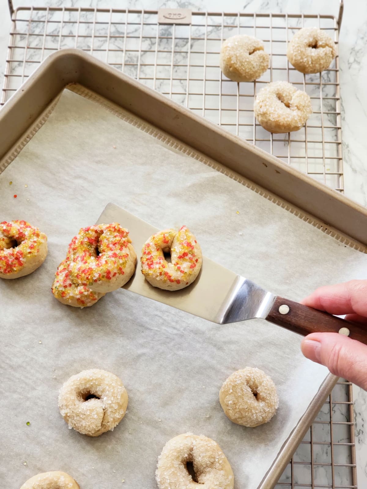 Hand holding a spatula has 2 cookies being moved to a cooling rack, from a gold colored baking sheet