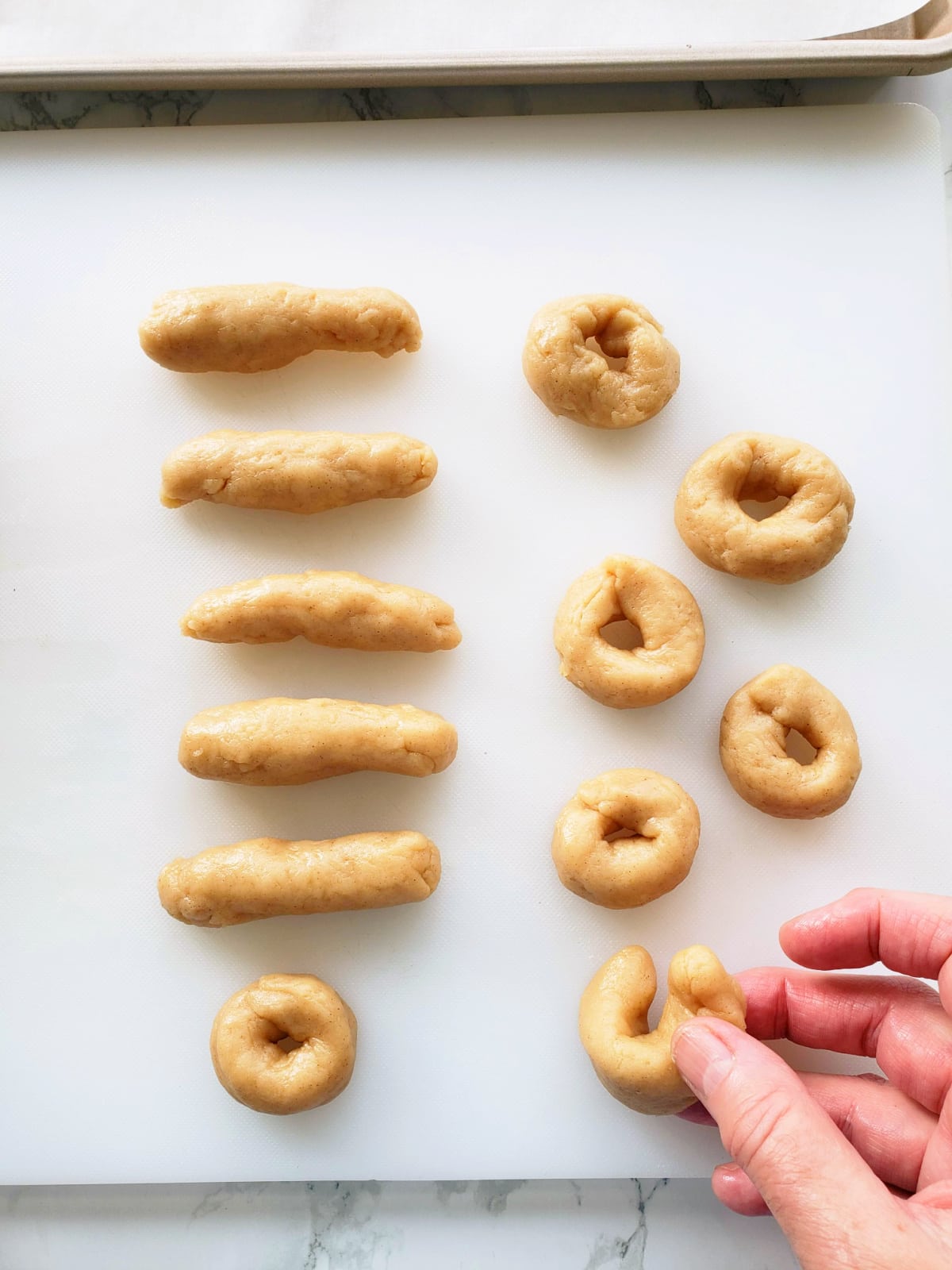 A hand forms a donut out of a dough cylinder on a white cutting board, with other dough cylinders on the board