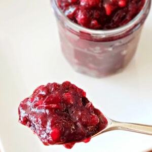 A spoon with cranberry sauce in foreground, a full jar of it behind, on a white background