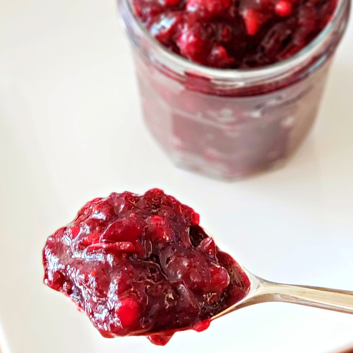 A spoon with cranberry sauce in foreground, a full jar of it behind, on a white background