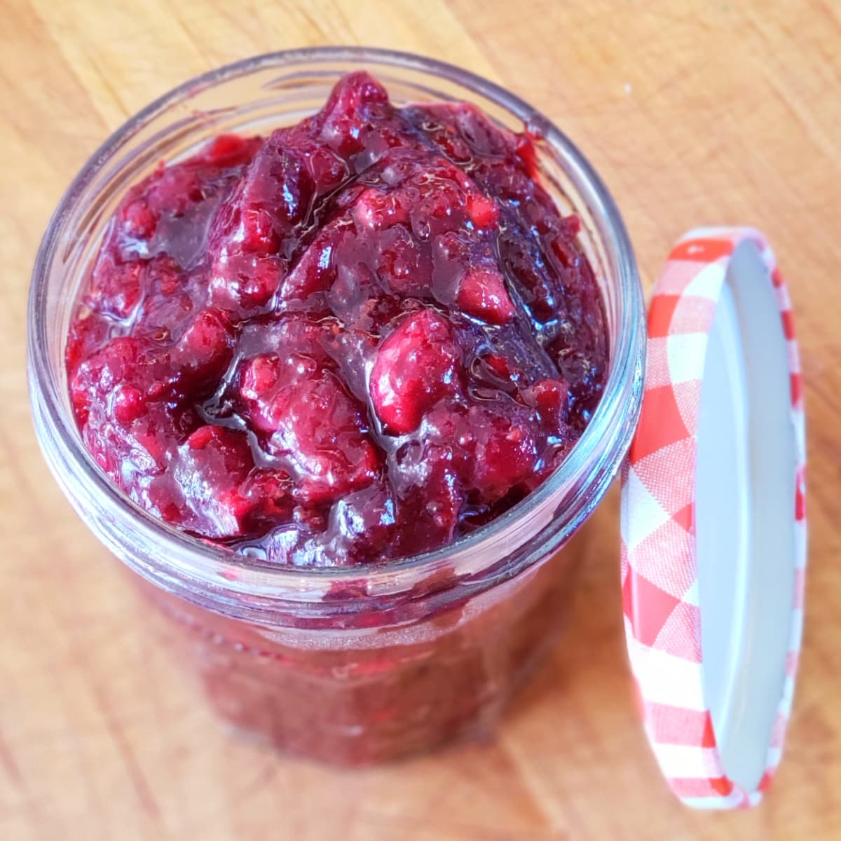 Skillet Cranberry Sauce with Port in a glass jar with a red gingham lid resting alongside it on a wooden board