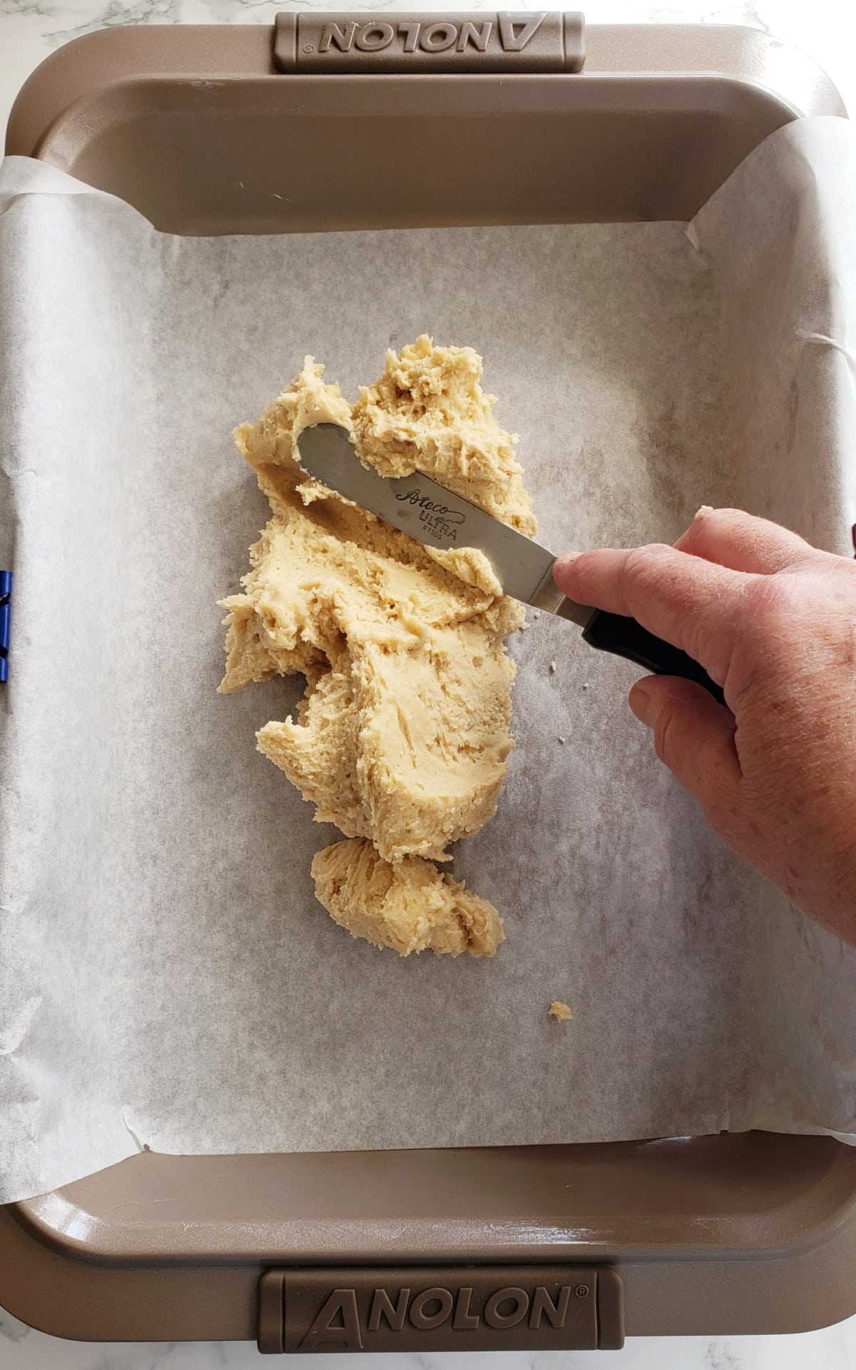 Hand holding an offset spatula spreads bottom dough into prepared baking dish