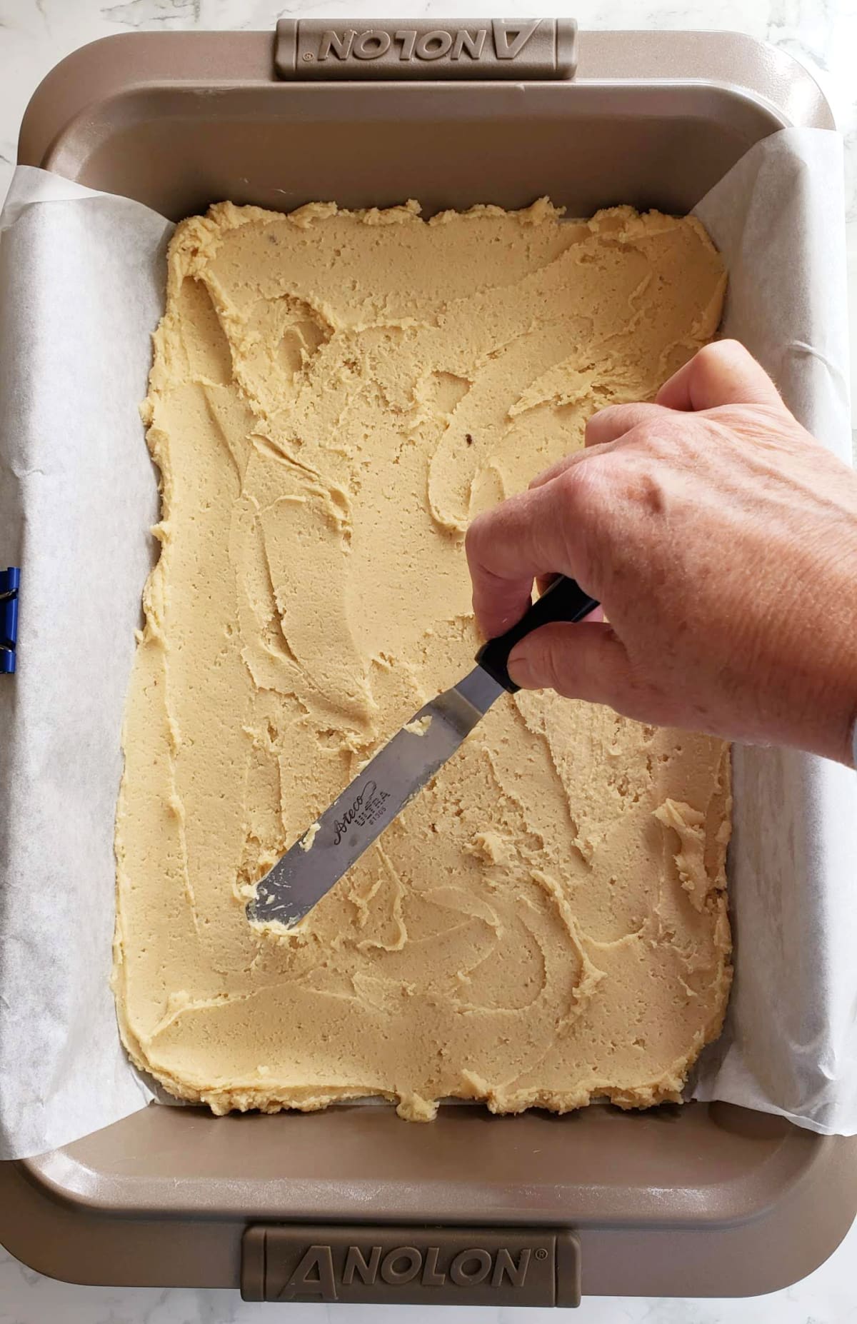 Hand holds a small offset spatula and smoothes the dough into the baking dish