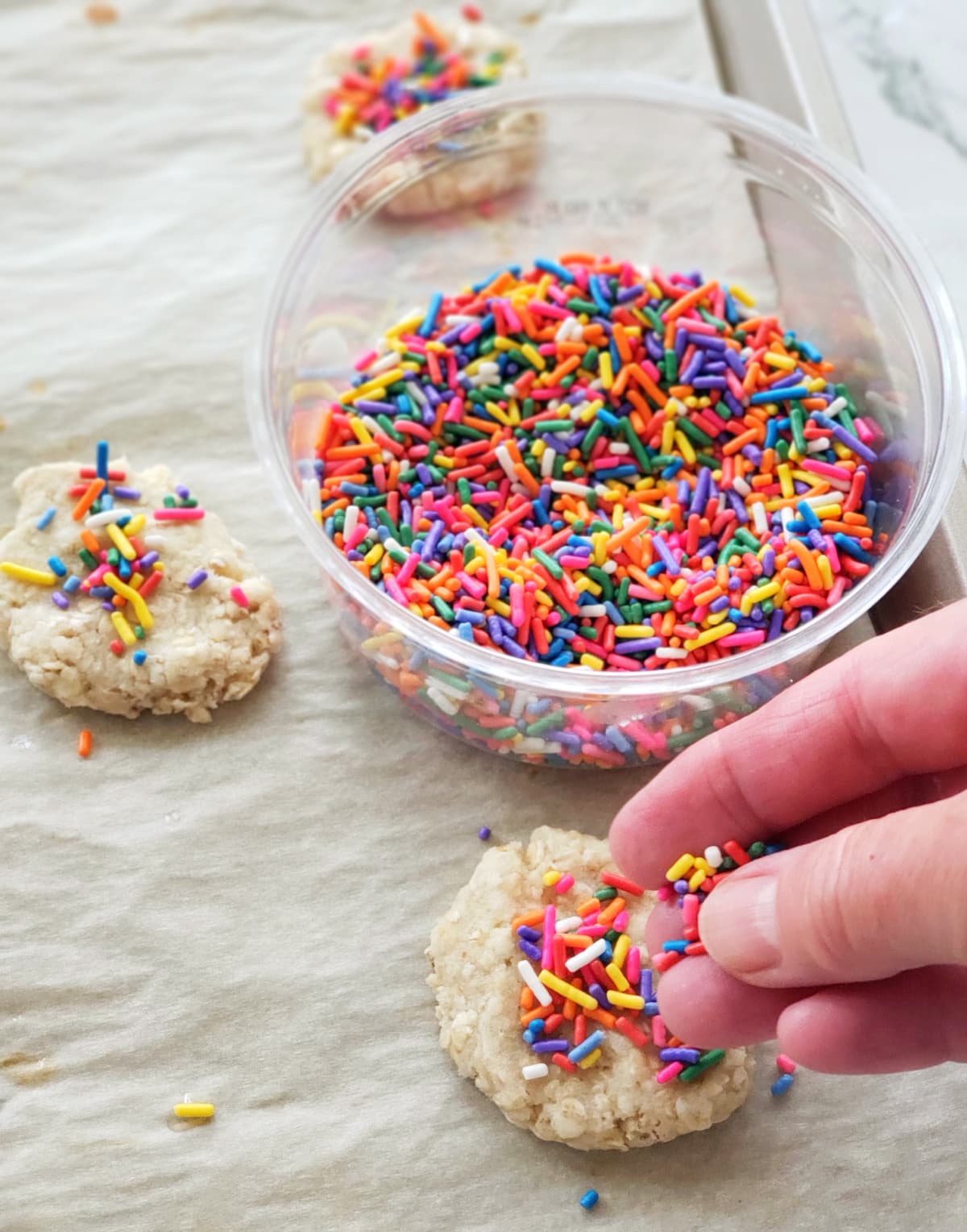 A hand adds sprinkles to the top of a cookie with a clear plastic container of sprinkles in the background of the cookies