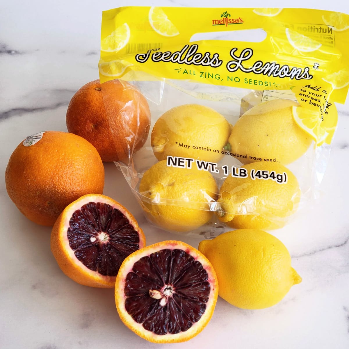 3 blood oranges, 1 cut in half to show the red interior, and a bag of lemons on a white countertop