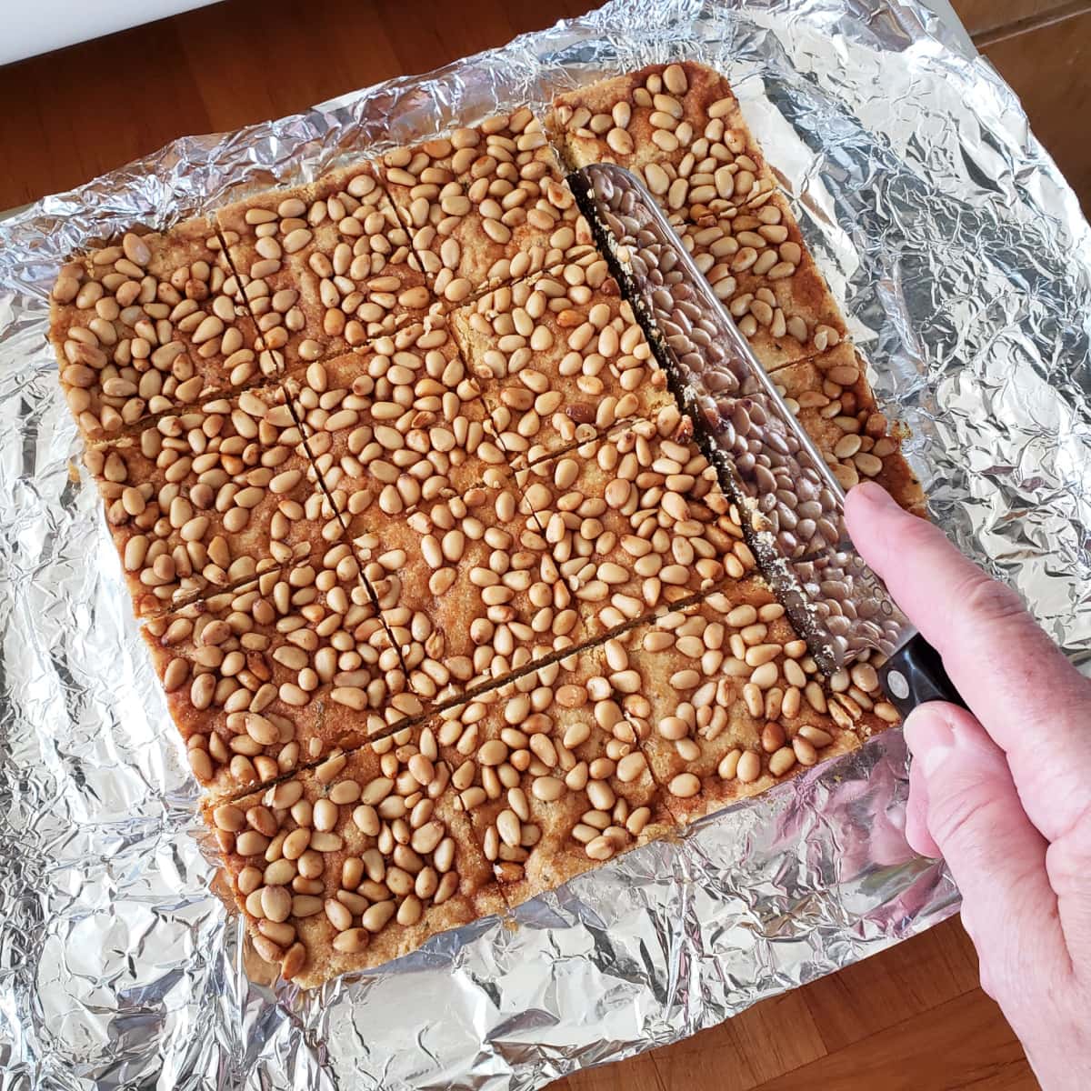 Hand holding large knife cuts pine nut cookies into squares, atop foil sheet resting on a wooden cutting board