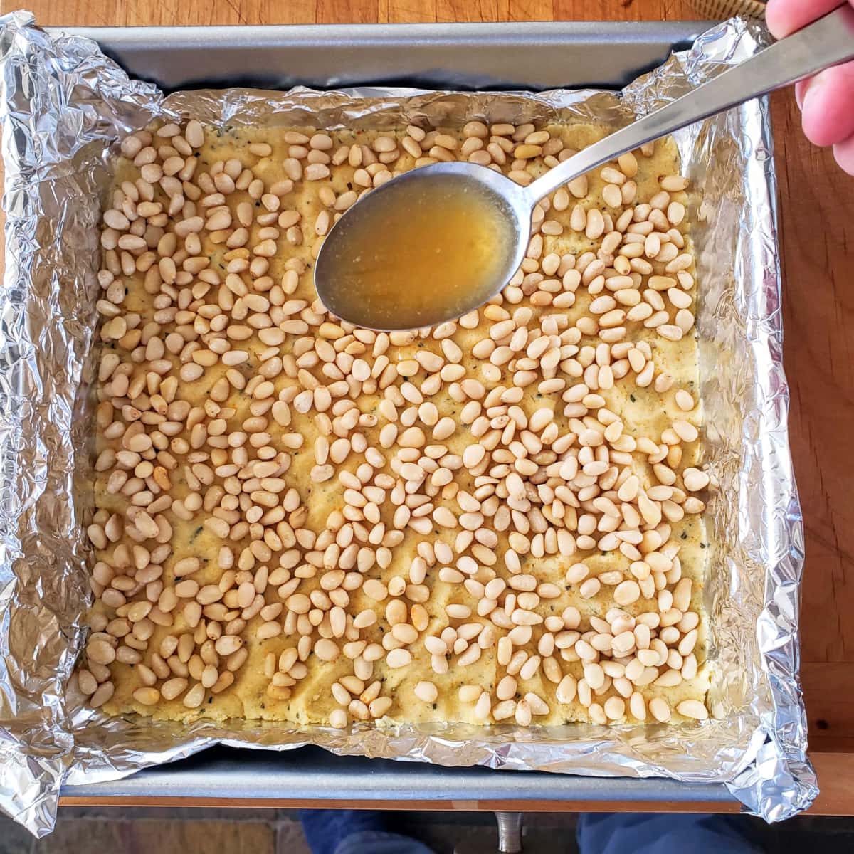 Hand holding a spoon drizzles butter honey over top of pine nuts in a silver baking dish