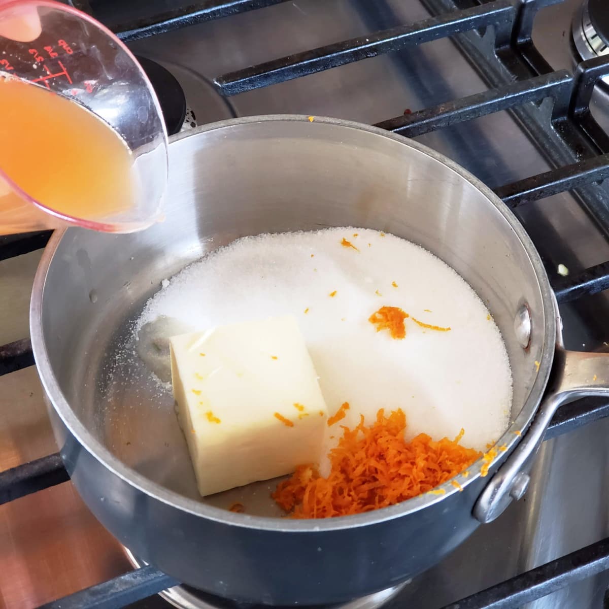 A hand pours orange juice from a measuring cup into a small silver sauce pan containing sugar, butter and orange zest
